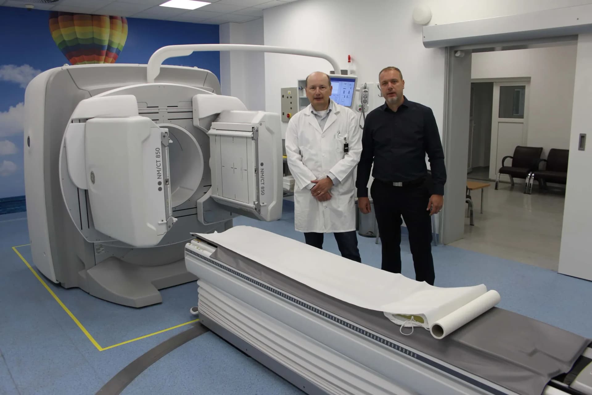 Dvaja muži, jeden v bielom plášti, stoja v miestnosti pri prístroji s označením NM/CT 850 a vyšetrovacím lôžkom