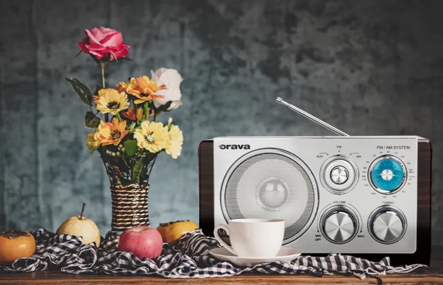 Retro rádio Orava s anténou a otočnými ovládačmi vedľa vázy s kvetmi, šálky a jabĺk