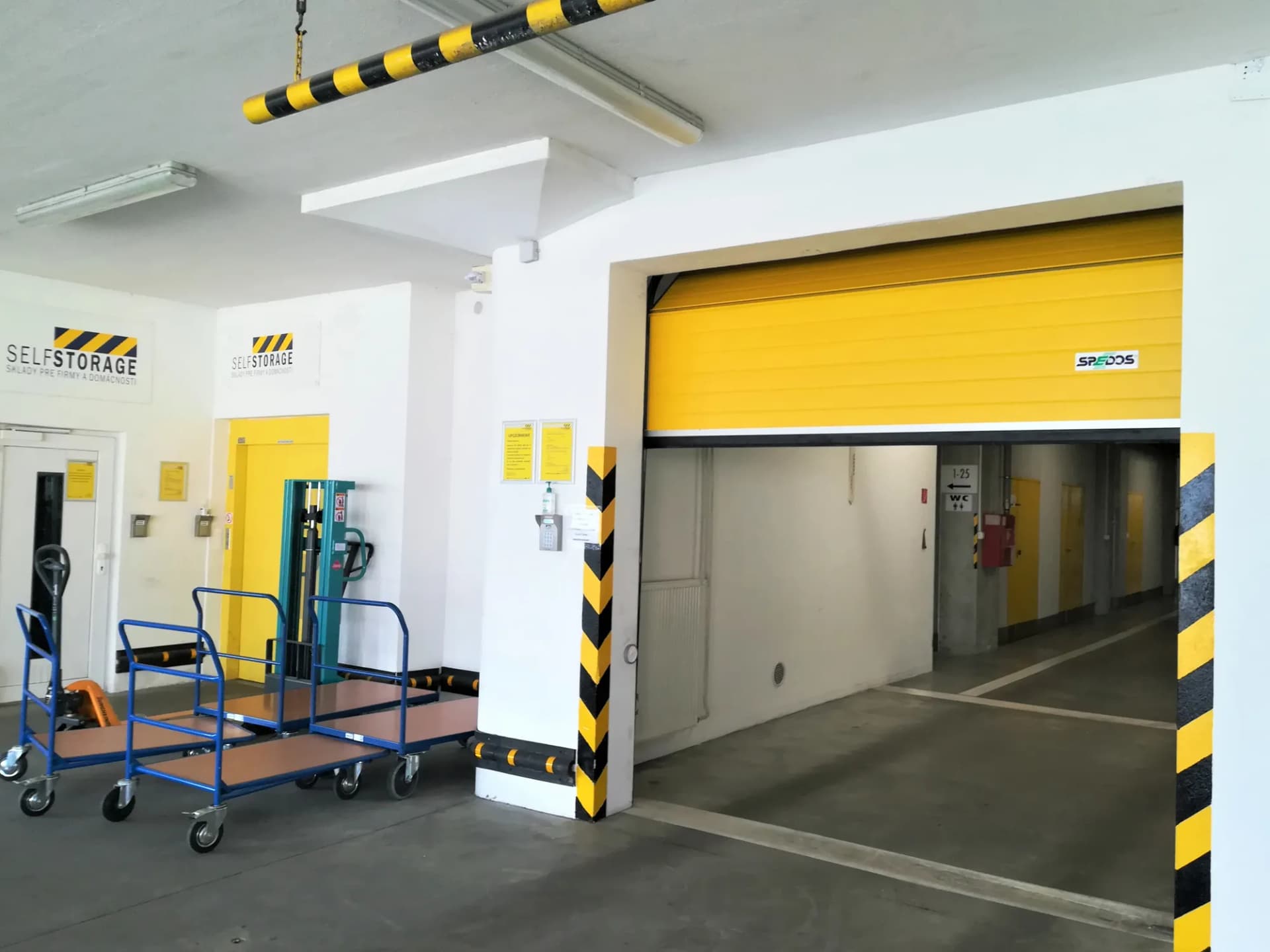 Vstupná hala skladu s modrými vozíkmi, paletovým vozíkom a žltou bránou do chodby s boxmi
